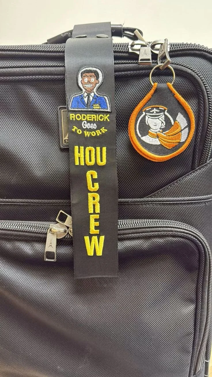 A black carry-on bag with two embroidered patches and a fabric tag. One patch features a cartoon woman in a graduation cap and gown, holding a diploma, with a white background and orange border. The other patch has a cartoon man in a blue suit with a yellow name tag that reads 'Roderick' and the words 'Good to Work' in cursive, along with the phrase 'HOU CREW' in bold yellow letters on the fabric tag.