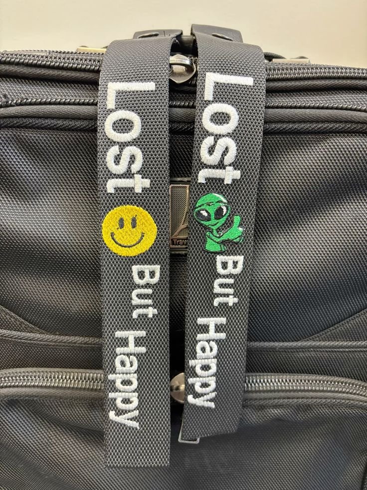 Close-up of a gray travel bag with two straps that have 'Lost' and 'Held' embroidered on them, one with a smiley face patch and one with an alien patch.