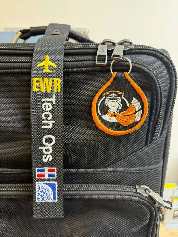 A black suitcase with a travel strap that has an airplane symbol, 'EWR,' and 'Tech Ops' embroidered on it, and a patch featuring a graduation cap, a face, and an orange scarf.