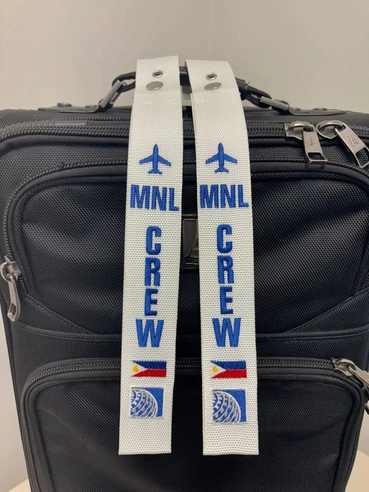 Two white luggage straps with blue text and logos, reading 'MNL CREW' and featuring Philippine flags and airline symbols, hanging from a black suitcase handle.