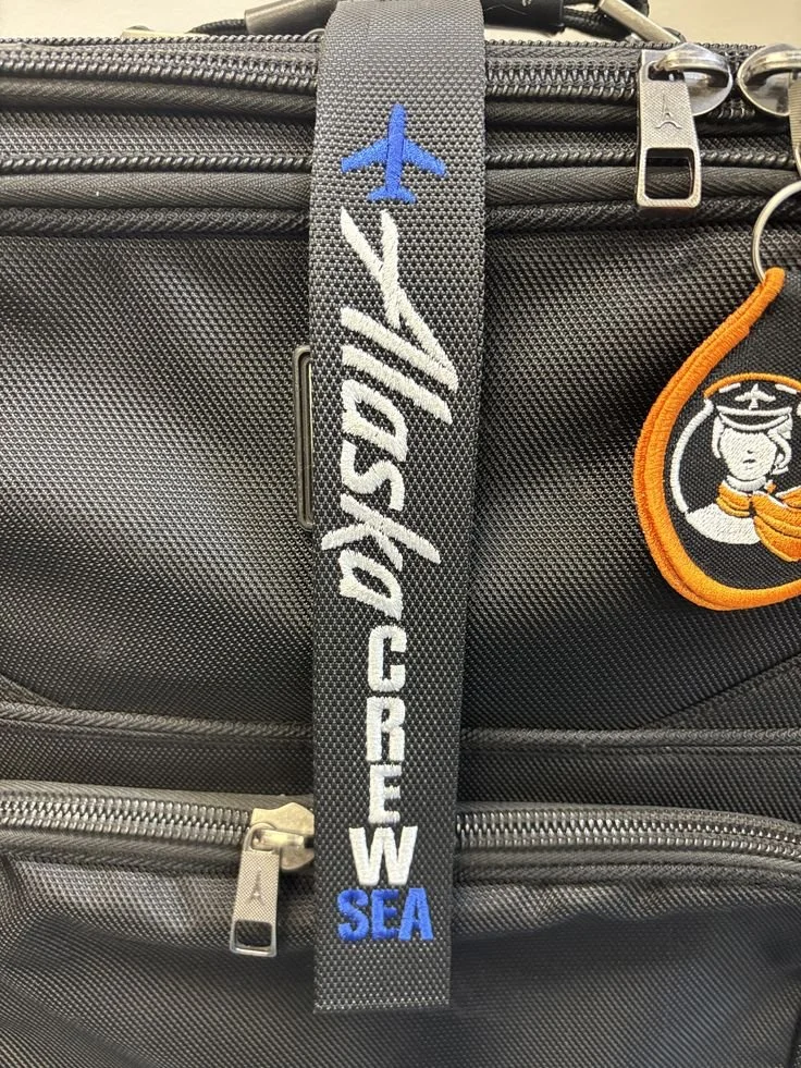 Close-up of a black and gray bag with a luggage tag featuring a blue airplane and the words 'ASHLEY CRAW SEA' embroidered on a strap.
