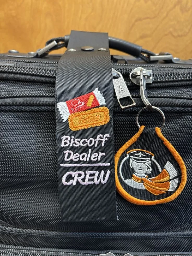 A close-up of a black travel bag with a zipper, featuring an embroidered patch of the cartoon character Snoopy dressed as a pilot, a logo patch reading 'Biscoff Dealership CREW', and a luggage strap.