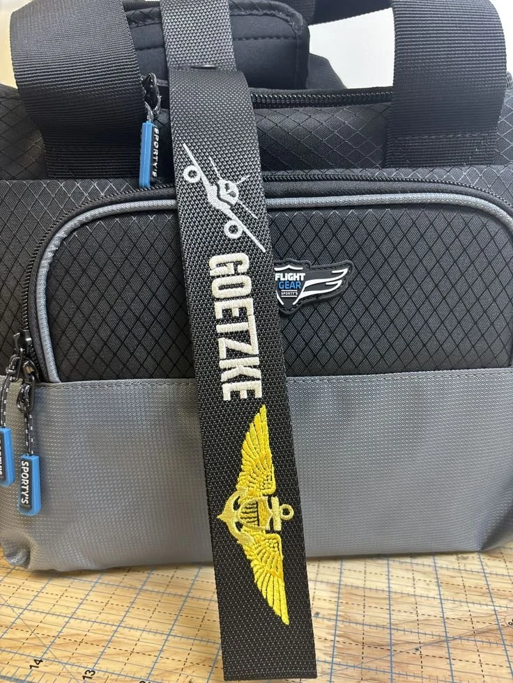 A black and gray airline travel bag with various zippers, tags, and patches, placed on a cutting mat. A lanyard with the text 'GUEZKE' and an embroidered yellow eagle with wings spread, along with a skull and wings logo, hangs over the bag.