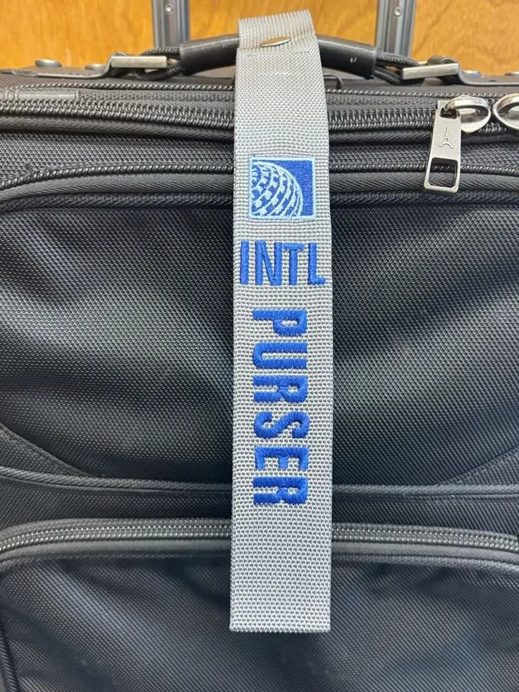 Close-up of a gray luggage strap with blue embroidered text 'INTL PURSER' and a globe logo, attached to a black wheeled suitcase.
