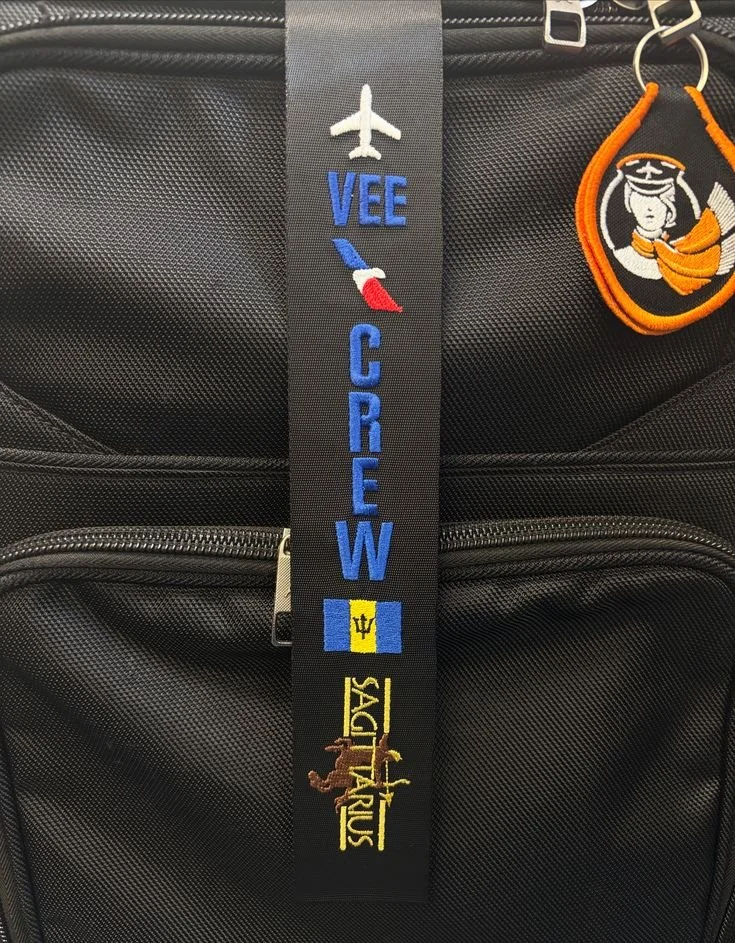 Close-up view of a luggage with a vertical strap that has embroidery of an airplane, the words "VEE CREW," and symbols representing France, the Vatican, and the Sacra Trinus logo. There is also a luggage tag with a cartoon character of a sailor.
