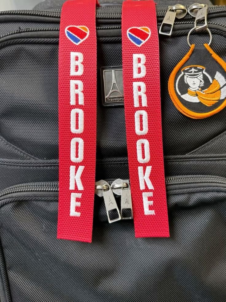 Close-up of a black travel bag with two red straps that have 'BROOKE' written on them and a heart with rainbow colors. There is also an orange and black embroidered keychain with a sailor girl logo attached to the bag.