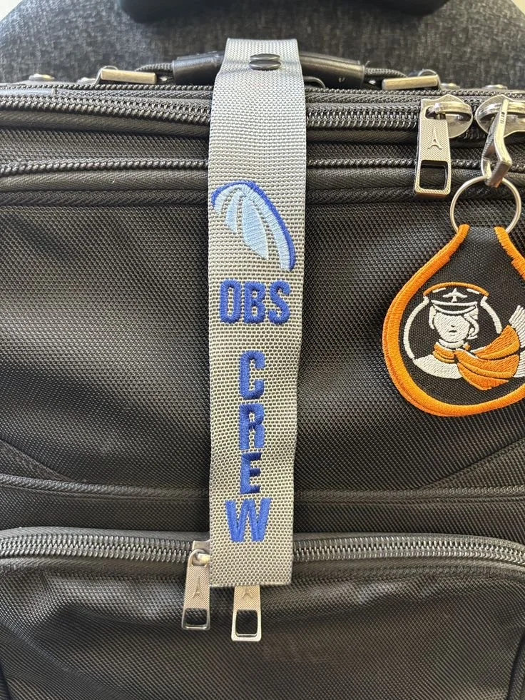 Close-up of a black travel bag with silver zippers, a gray strap with blue embroidery reading "OBS CREW" and a logo, and a keychain with a character in a captain's hat and orange scarf.