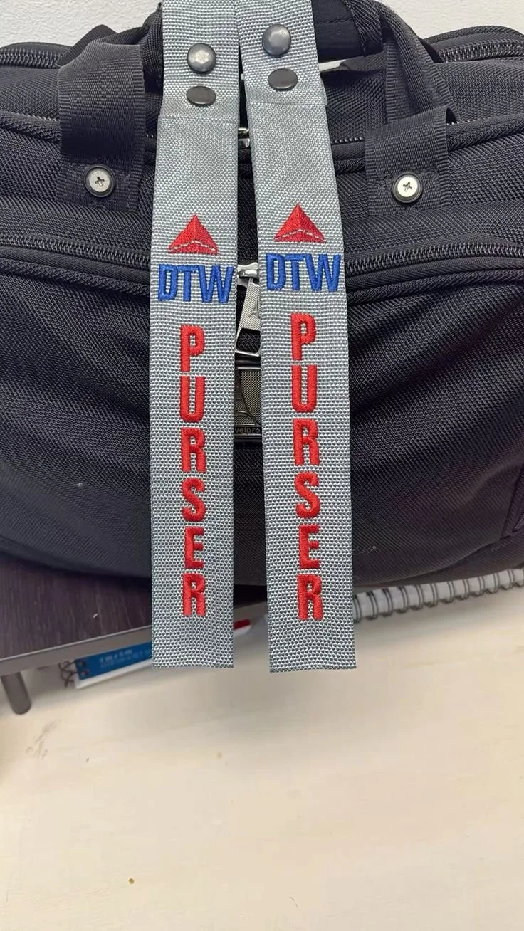 Close-up of two grey zipper pulls on a black bag, each embroidered with the words 'PURSER' in red and 'DTW' in blue, with a red triangle logo above the letters.