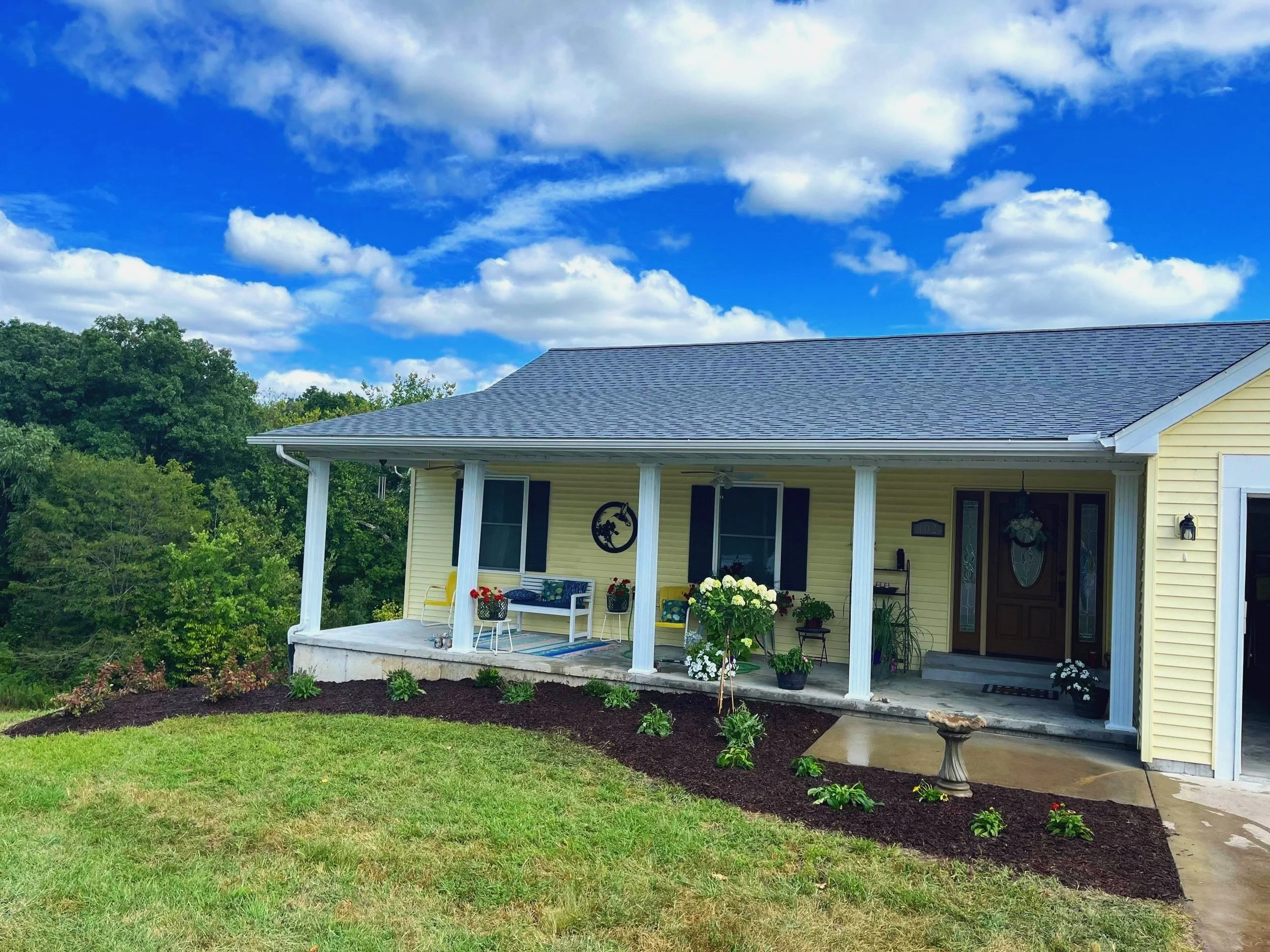 Yellow house with porch, black shutters, and garden with white flowers under blue sky with clouds.