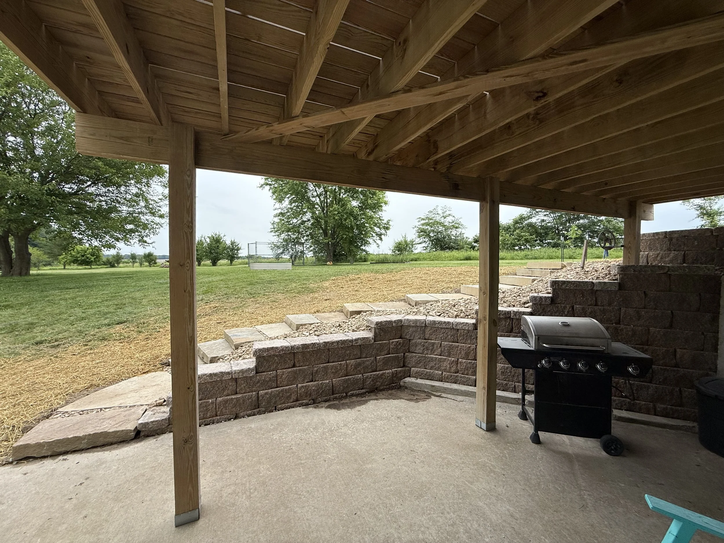 Covered outdoor patio with a black gas grill, brick retaining wall, and a grassy yard with trees in the background.