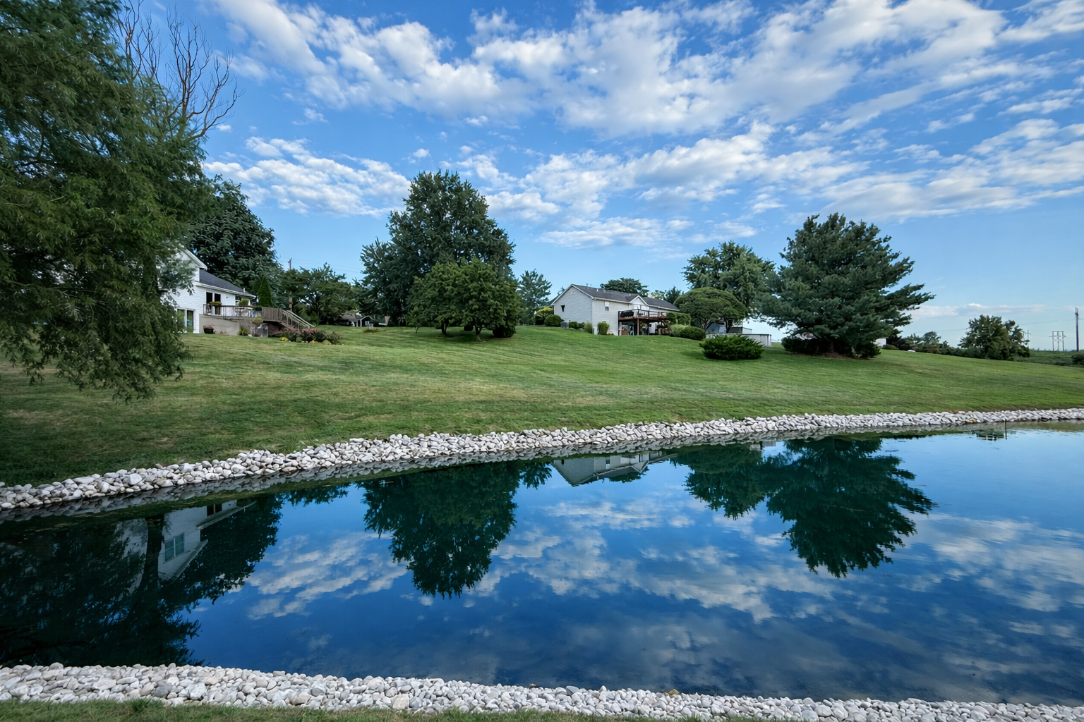 A peaceful backyard view featuring a pond with a rocky border, a grassy hill with several trees, and white residential houses under a partly cloudy sky.