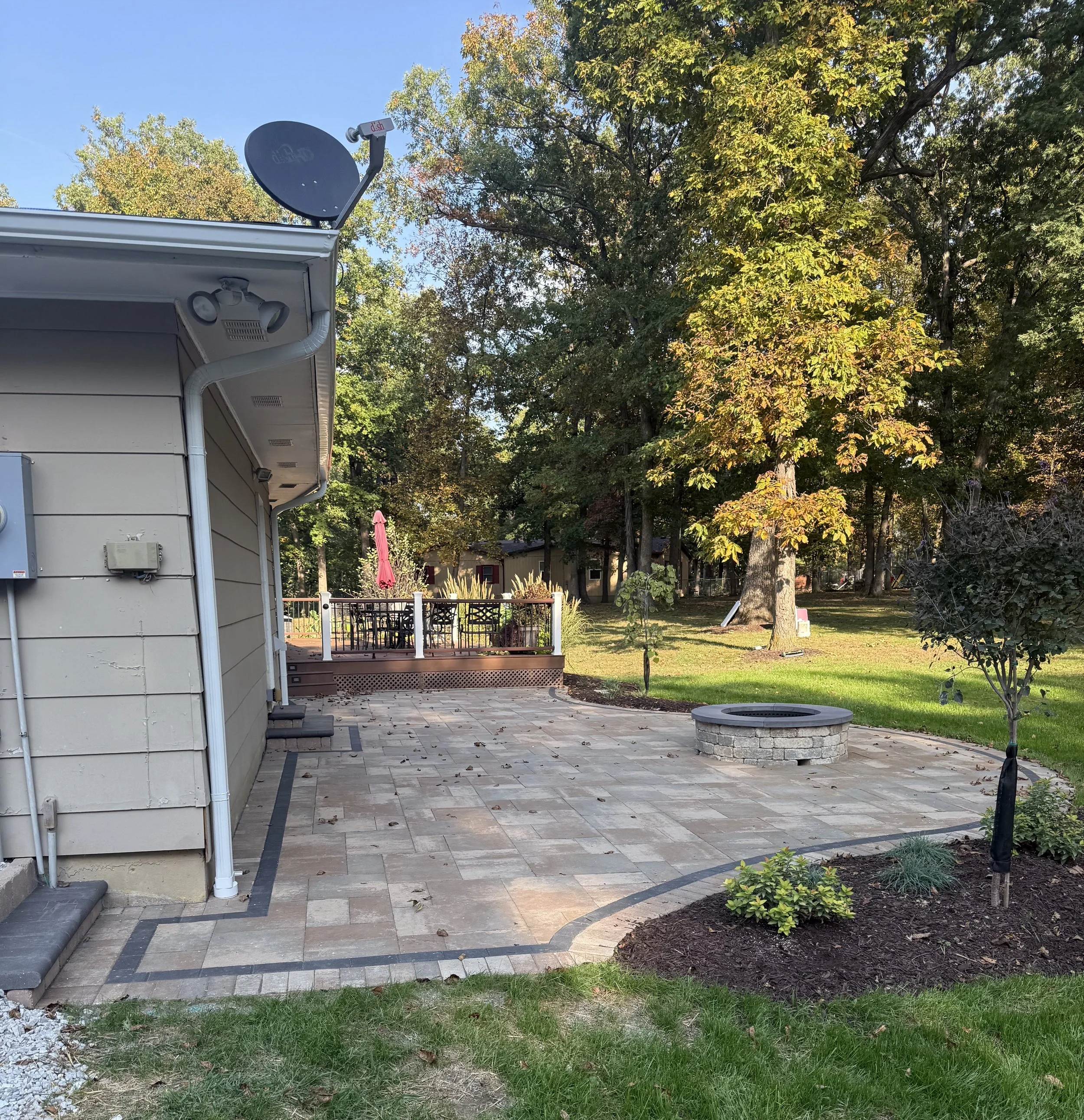 Backyard with stone patio, fire pit, and large trees with autumn-colored leaves.