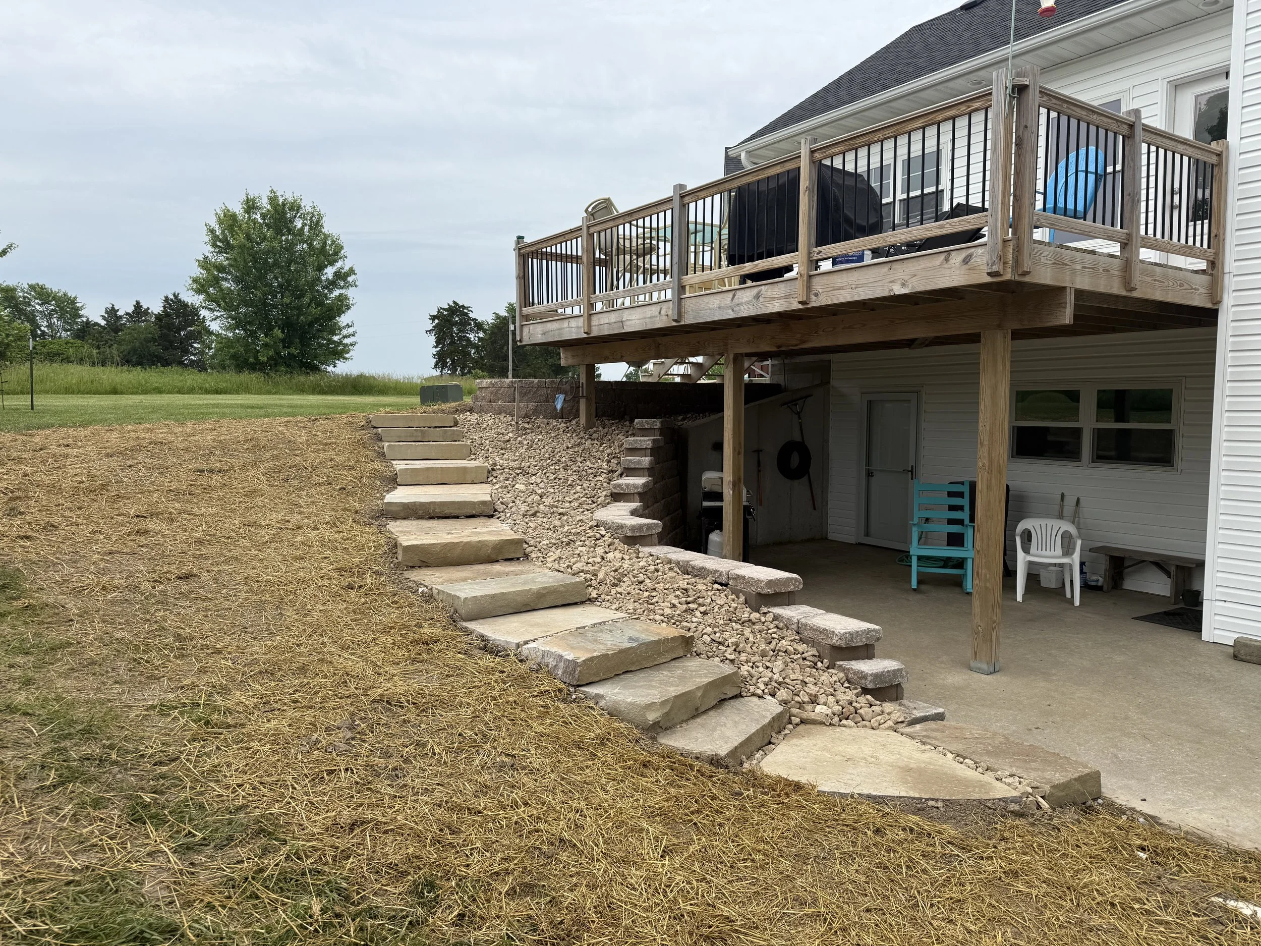 Backyard with wooden staircase leading from upper deck to ground level, with some chairs and equipment under the deck, and a grassy area on the left.