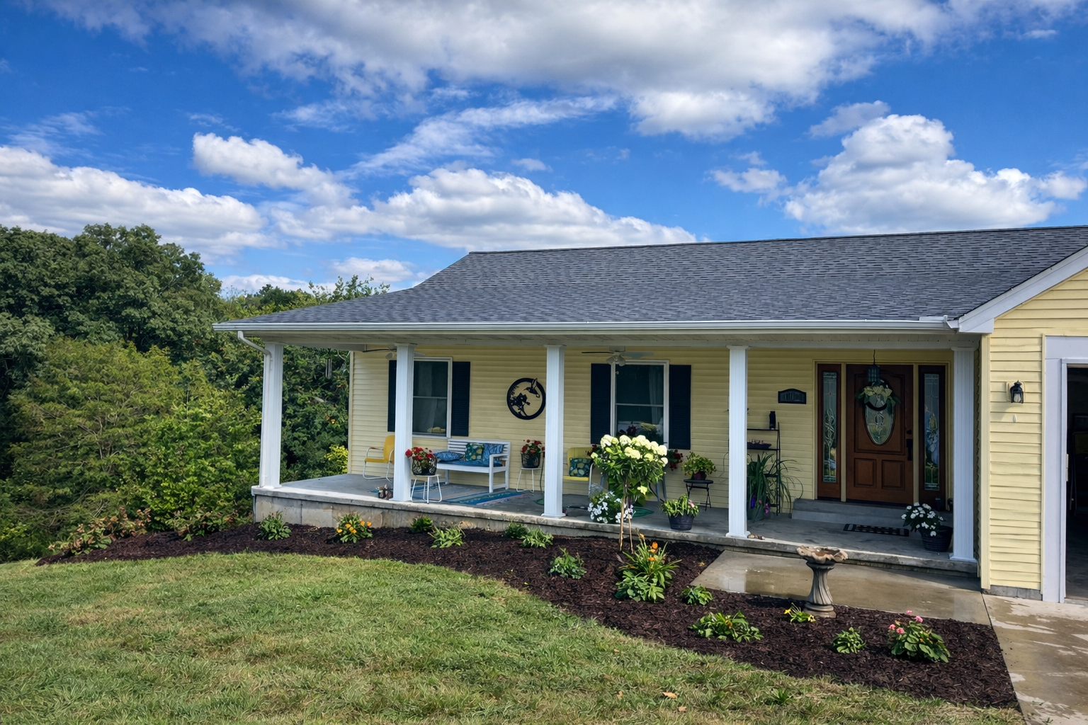 Yellow house with a porch decorated with flowers and outdoor furniture, set against a green, tree-filled background and a partly cloudy sky.