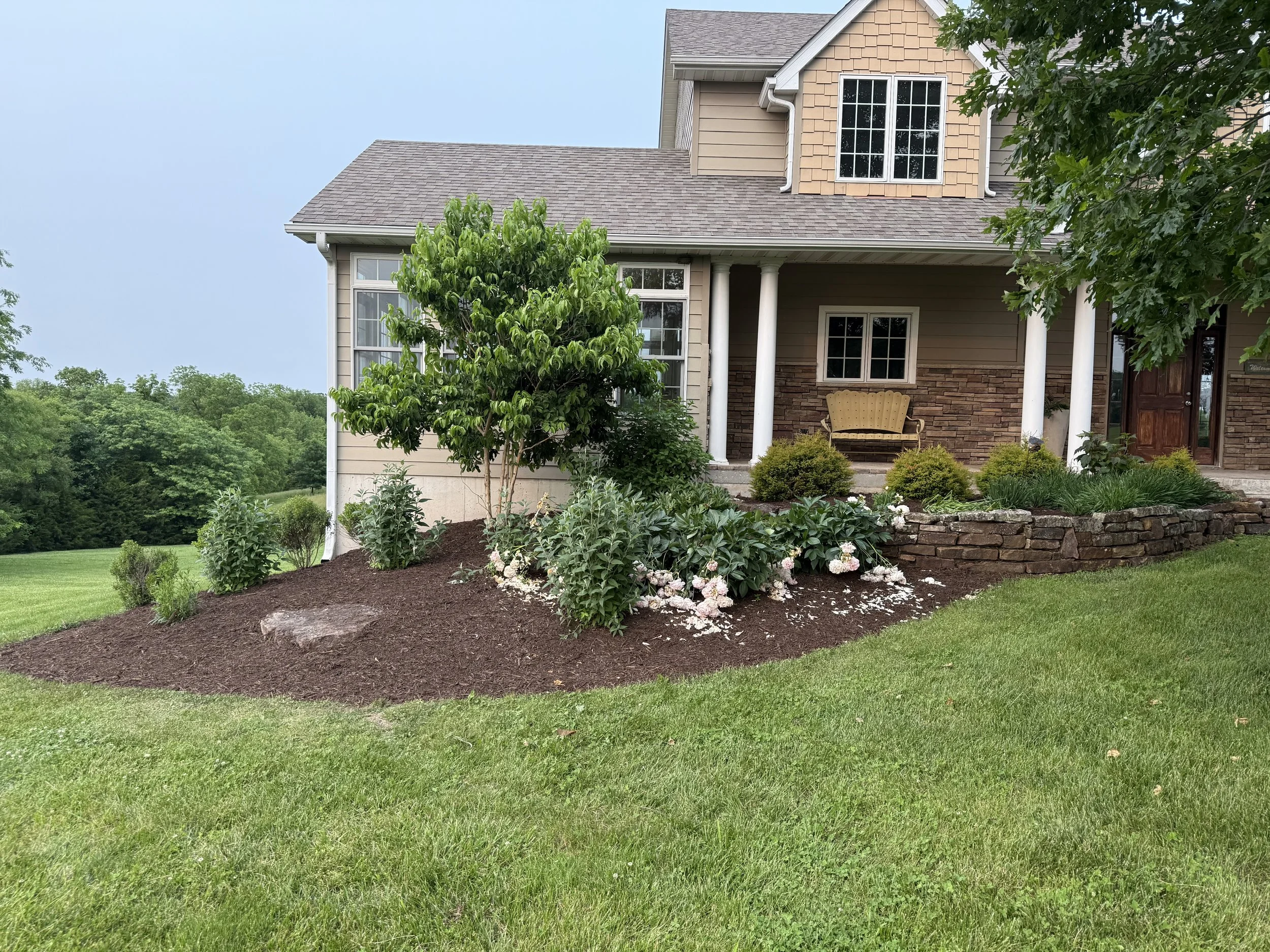 Front yard garden bed with bushes, flowers, and a small tree in front of a house with a porch and bench, surrounded by green grass and trees.