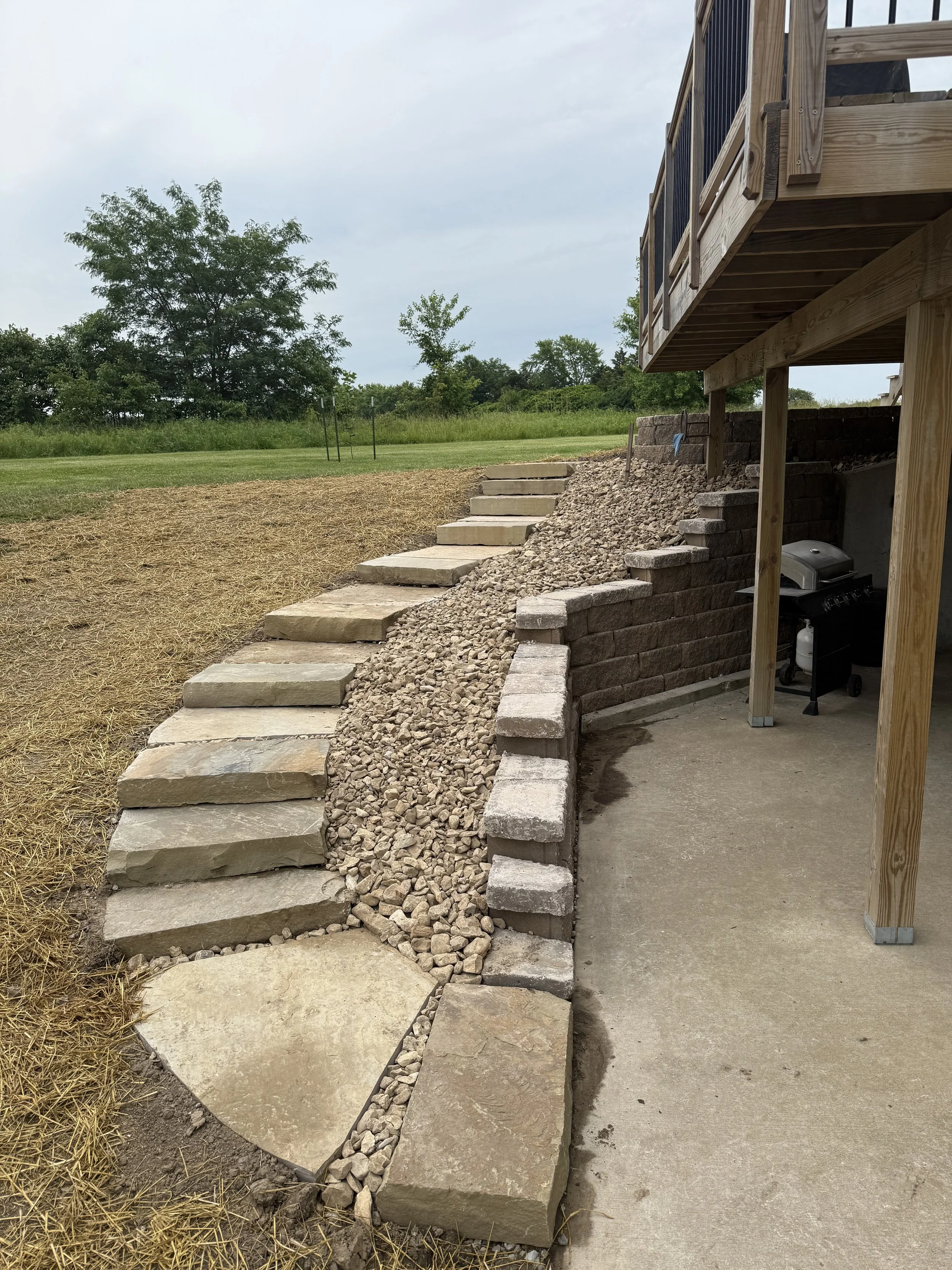 Stone steps and retaining wall leading from a grassy backyard to a higher level, next to a wooden deck with barbecue grill underneath, on concrete patio.