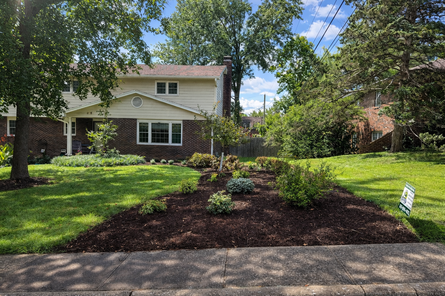 A residential front yard with a garden bed filled with plants, shrubs, and flowers, adjacent to a concrete sidewalk and lush green lawn, with a house and trees in the background under a partly cloudy sky.