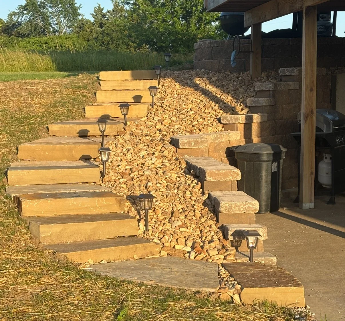 Outdoor staircase with large wooden steps and a central rock-filled drainage channel, illuminated by small black lamps, leading up to a garden area with grass and trees.