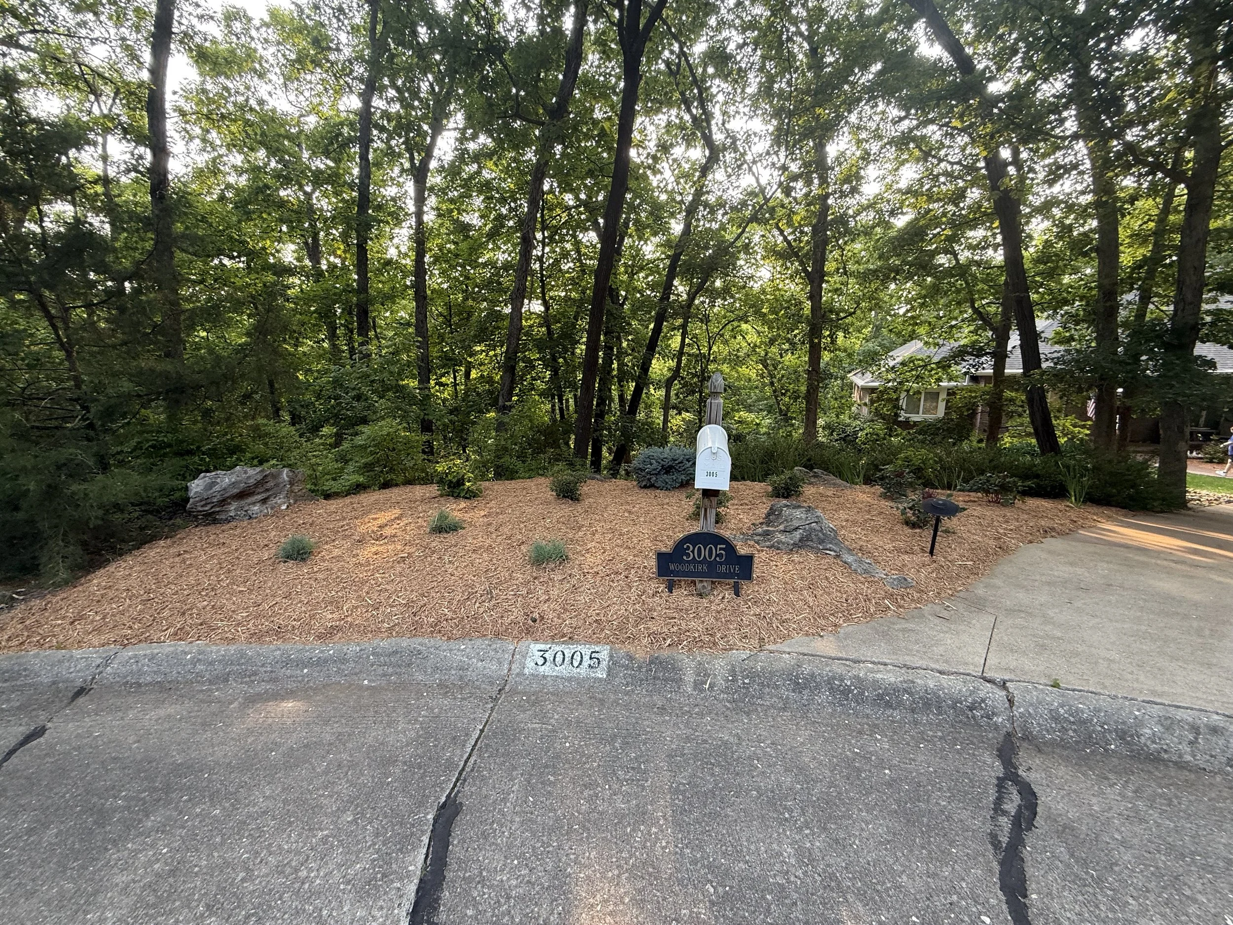 Residential house address marker, mail collection box, and landscaped yard at 3005 Woodkirk Drive, with trees in the background.