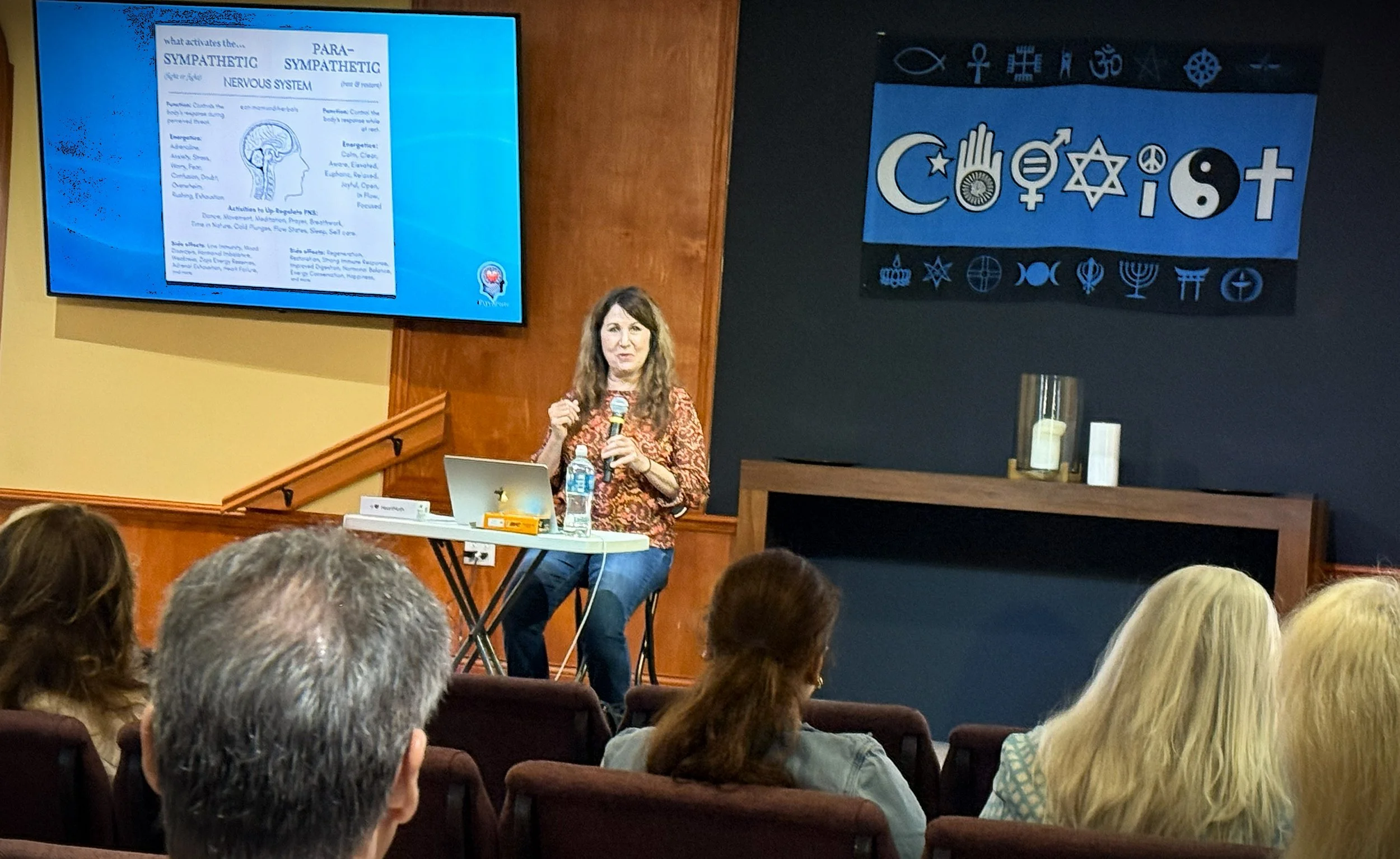 Speaker Heather Whitefield sits at a small table with a microphone during a presentation on the nervous system at Unity of Daytona Beach. A slide showing sympathetic and parasympathetic nervous system diagrams is projected on the screen beside her. A