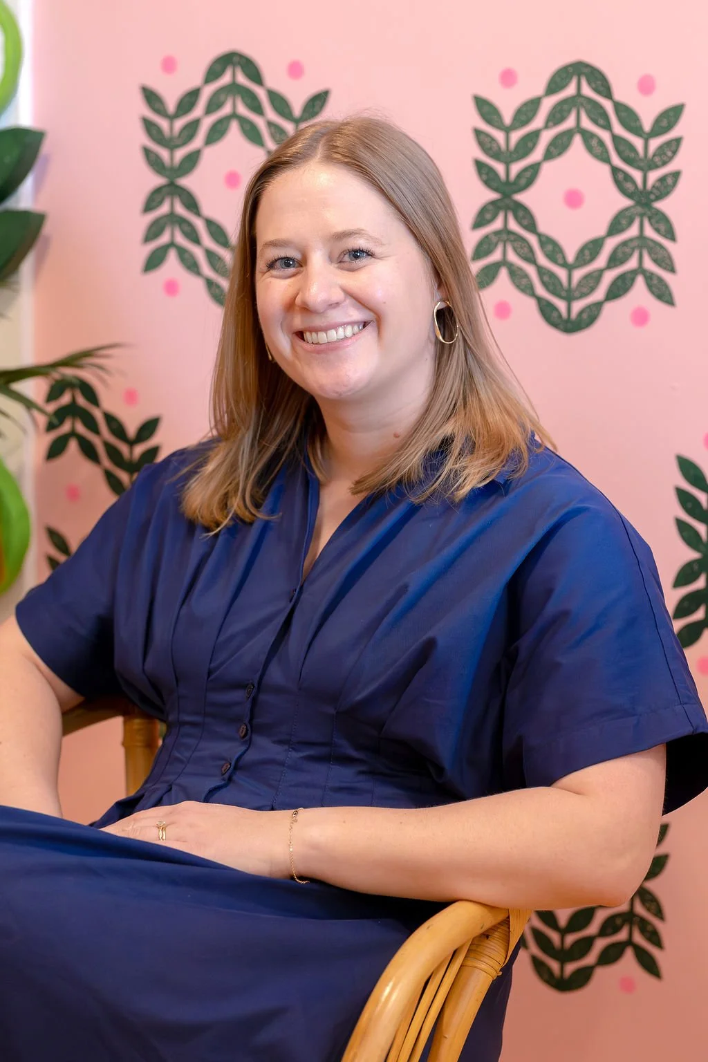 A woman with shoulder-length light brown hair smiling, wearing a navy blue dress with button details and hoop earrings, sitting in front of a pink wall with leafy black patterns and pink dots.