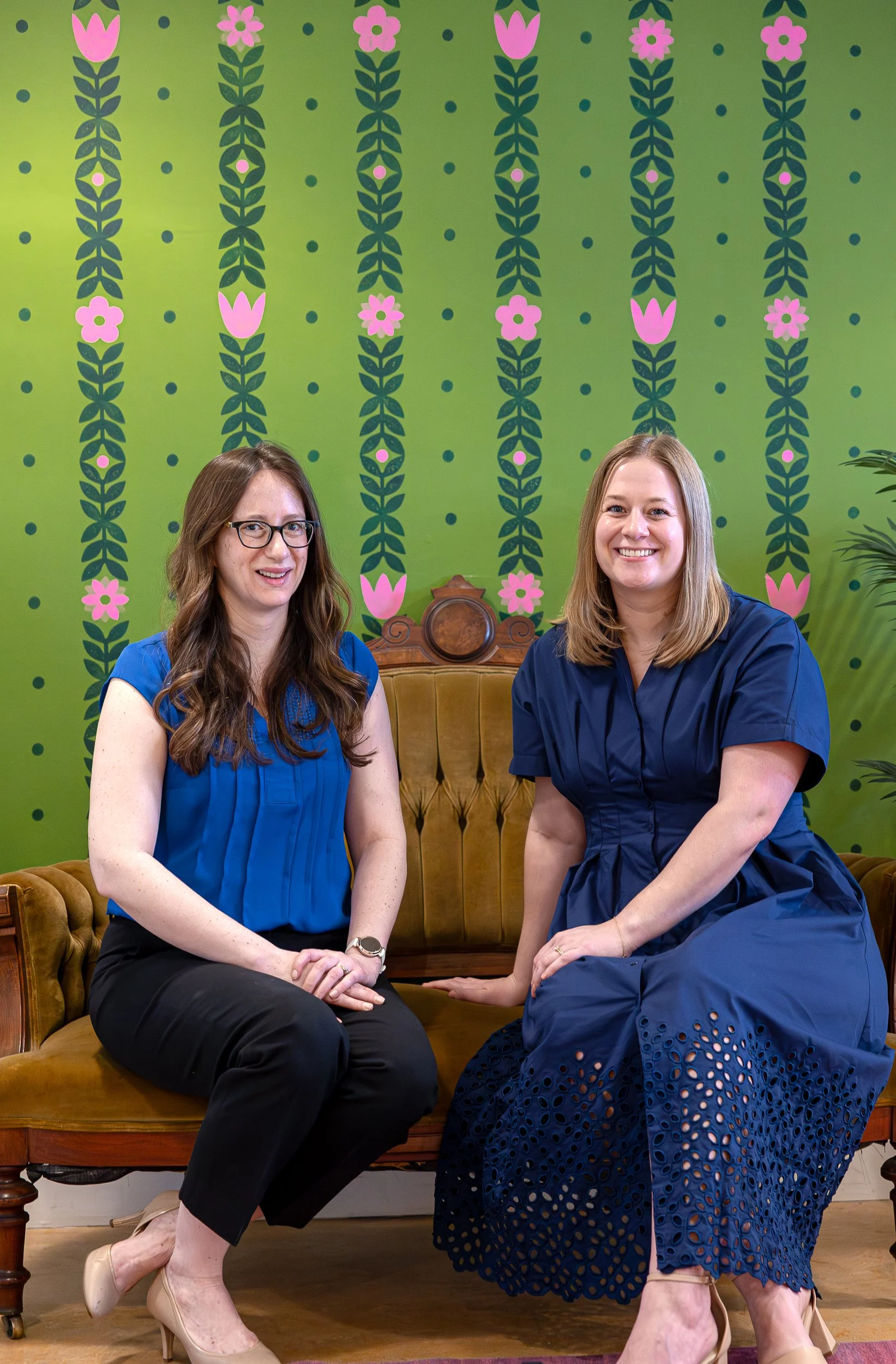 Two women sitting on a vintage wooden and velvet sofa in front of a green floral patterned wallpaper. The woman on the left has long brown hair, glasses, and is wearing a blue blouse with black pants and beige heels. The woman on the right has shoulder-length blond hair, a blue dress with eyelet details, and beige heels. They are smiling at the camera.