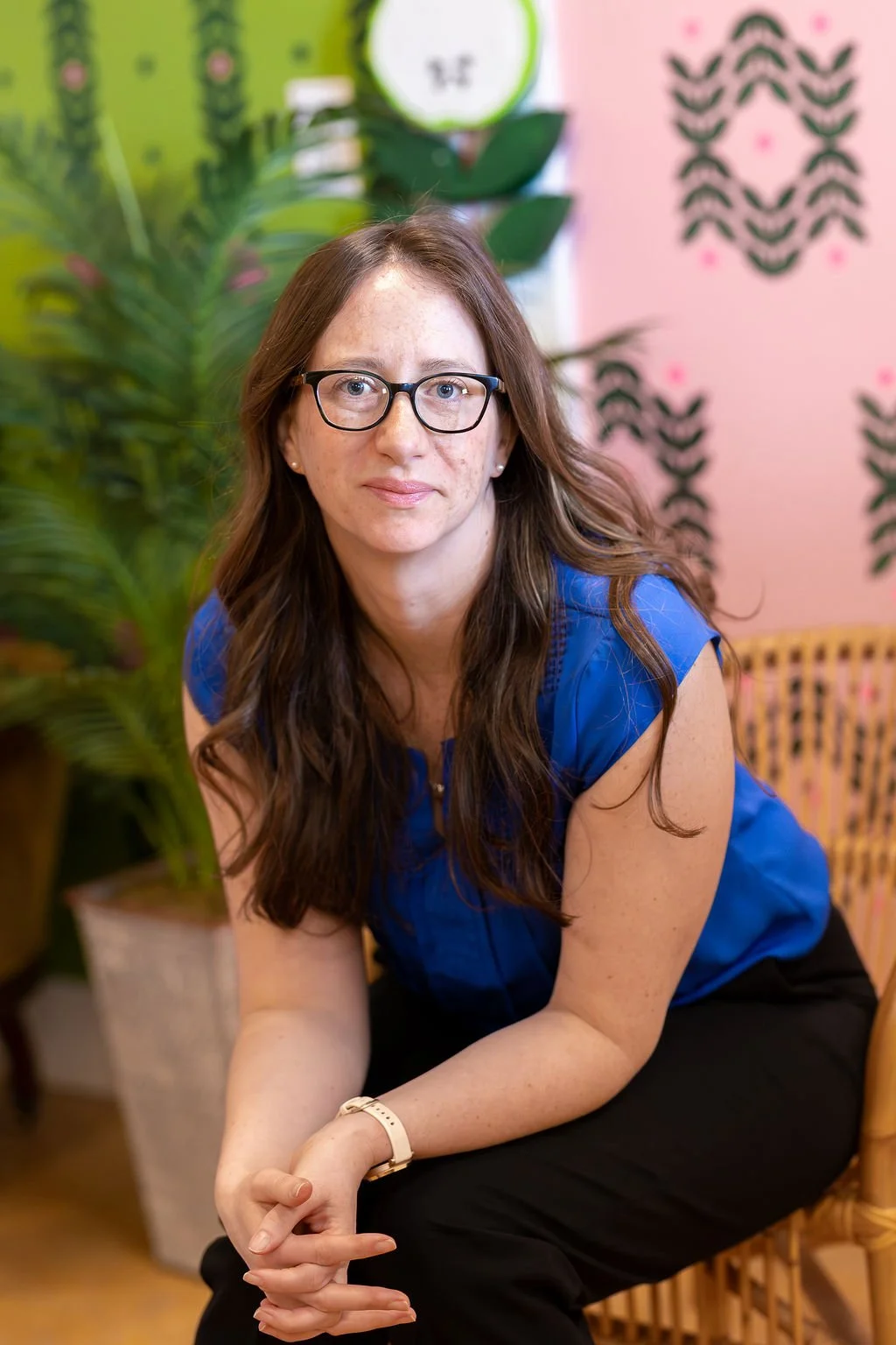 A woman with long brown hair, glasses, and a blue blouse, sitting on a wicker chair in a colorful room with green plants and pink walls with black and pink patterns.