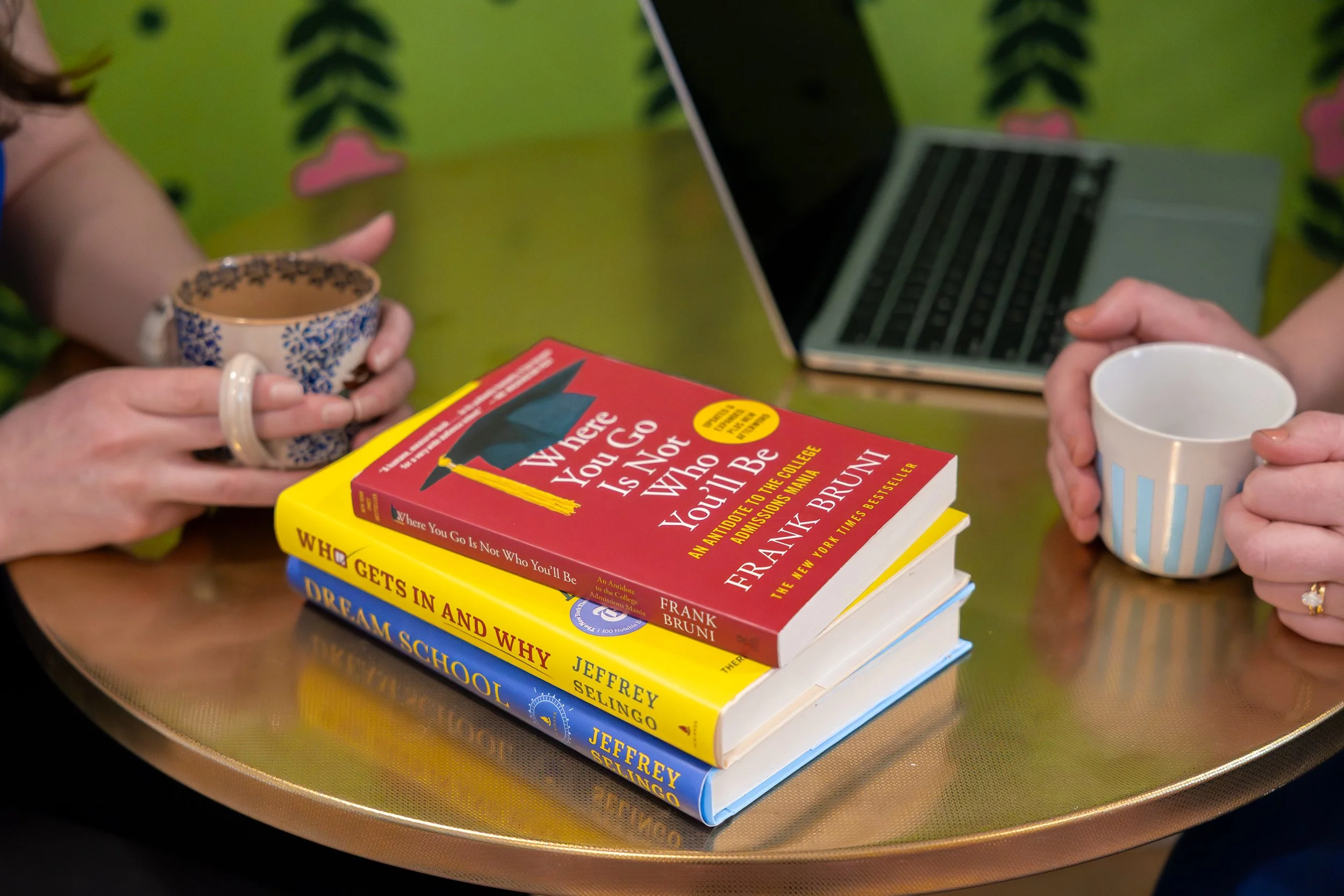 Two people sitting at a table with a laptop and a stack of three books, two cups of coffee or tea, and a gold-colored table. The top book is red titled 'Where You Go Is Not Who You Be' by Frank Bruni, the middle book is yellow, and the bottom book is blue.