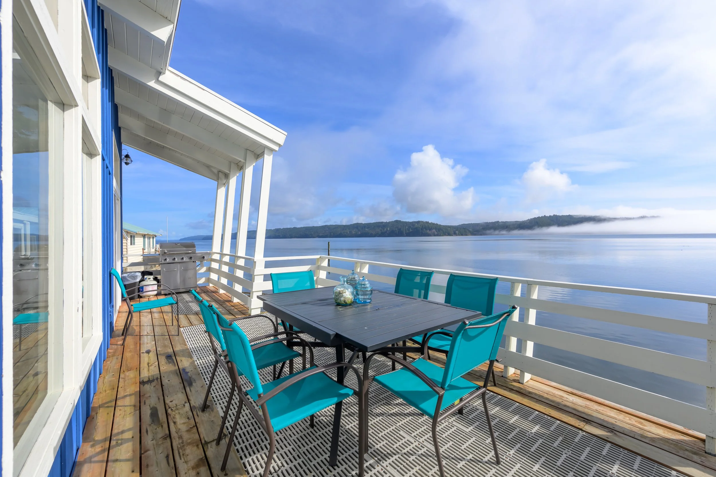 A balcony with a table and blue chairs overlooking a body of water and distant hills under a partly cloudy sky.