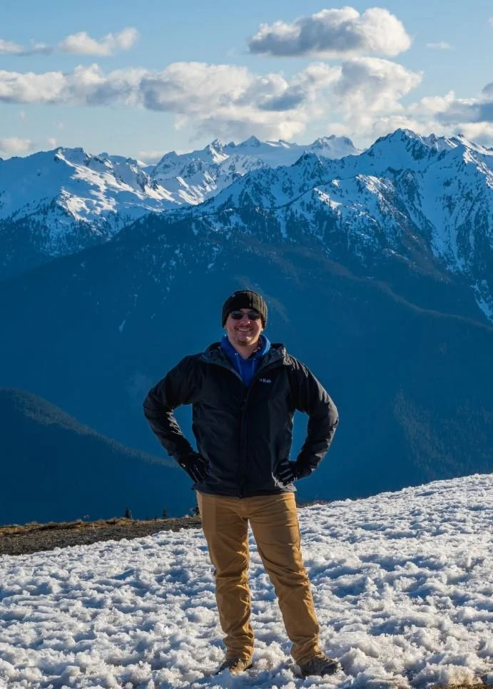 A man dressed in winter clothing, including a black jacket, tan pants, and a black beanie, stands smiling on a snowy mountain landscape with snow-capped peaks and blue skies in the background.