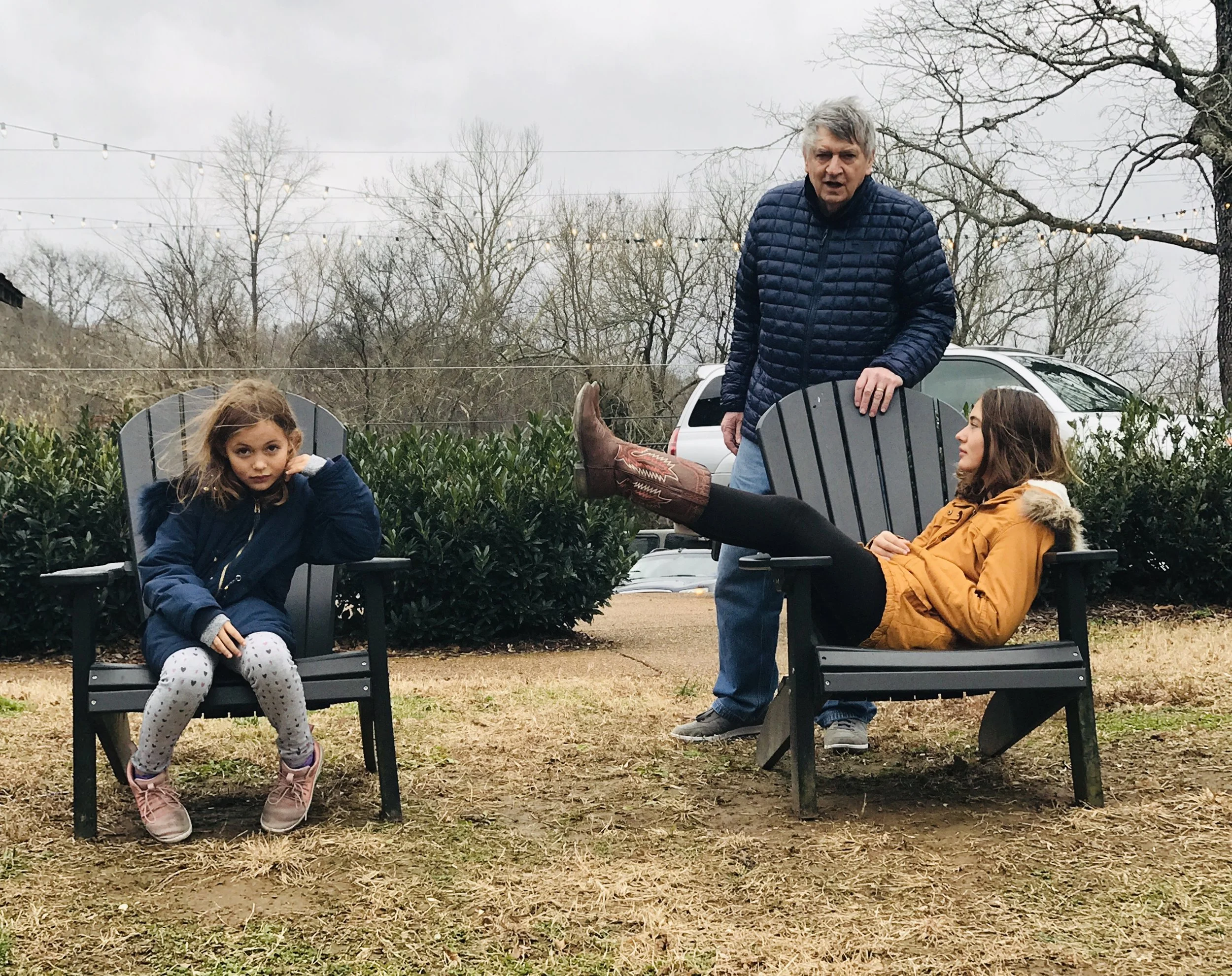 Three people sitting on black outdoor chairs in a yard with leafless trees and string lights. The person on the left is a young girl with brown hair in a blue jacket and gray leggings. The person standing in the middle is an older man with gray hair 