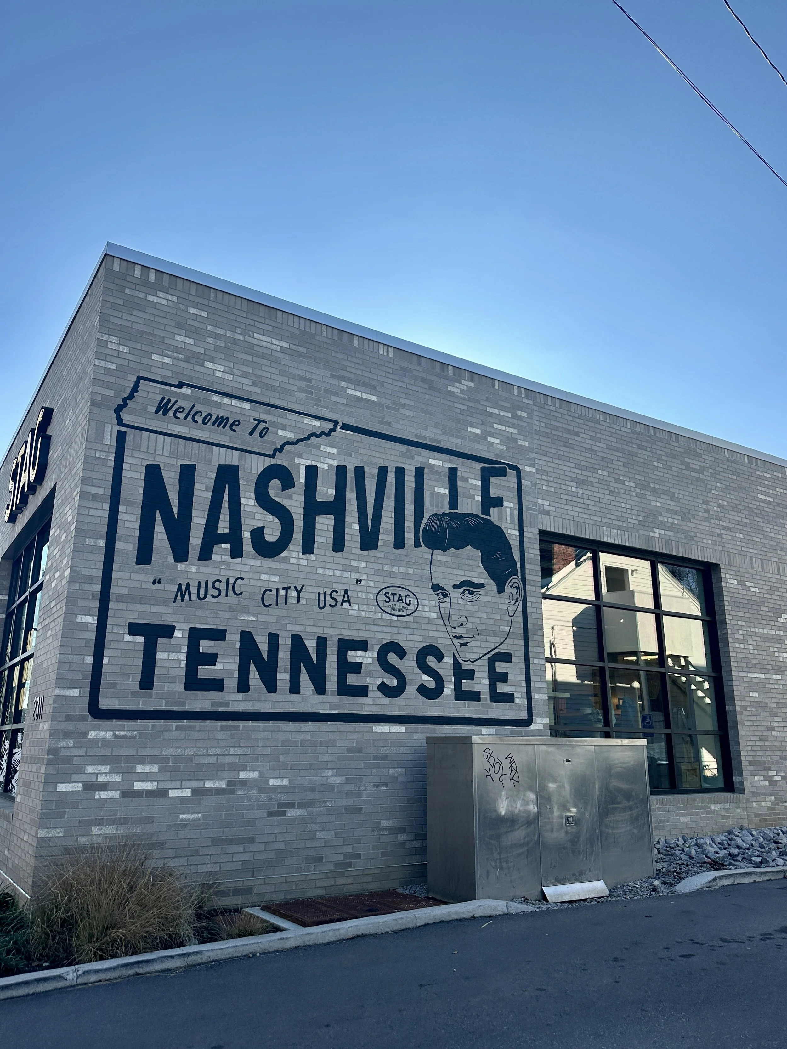 Street view of a building with a mural that reads "Welcome to Nashville, Music City USA." The mural includes a vintage black-and-white portrait of a man with a pompadour hairstyle. The building has large windows, a brick exterior, and a metal utility
