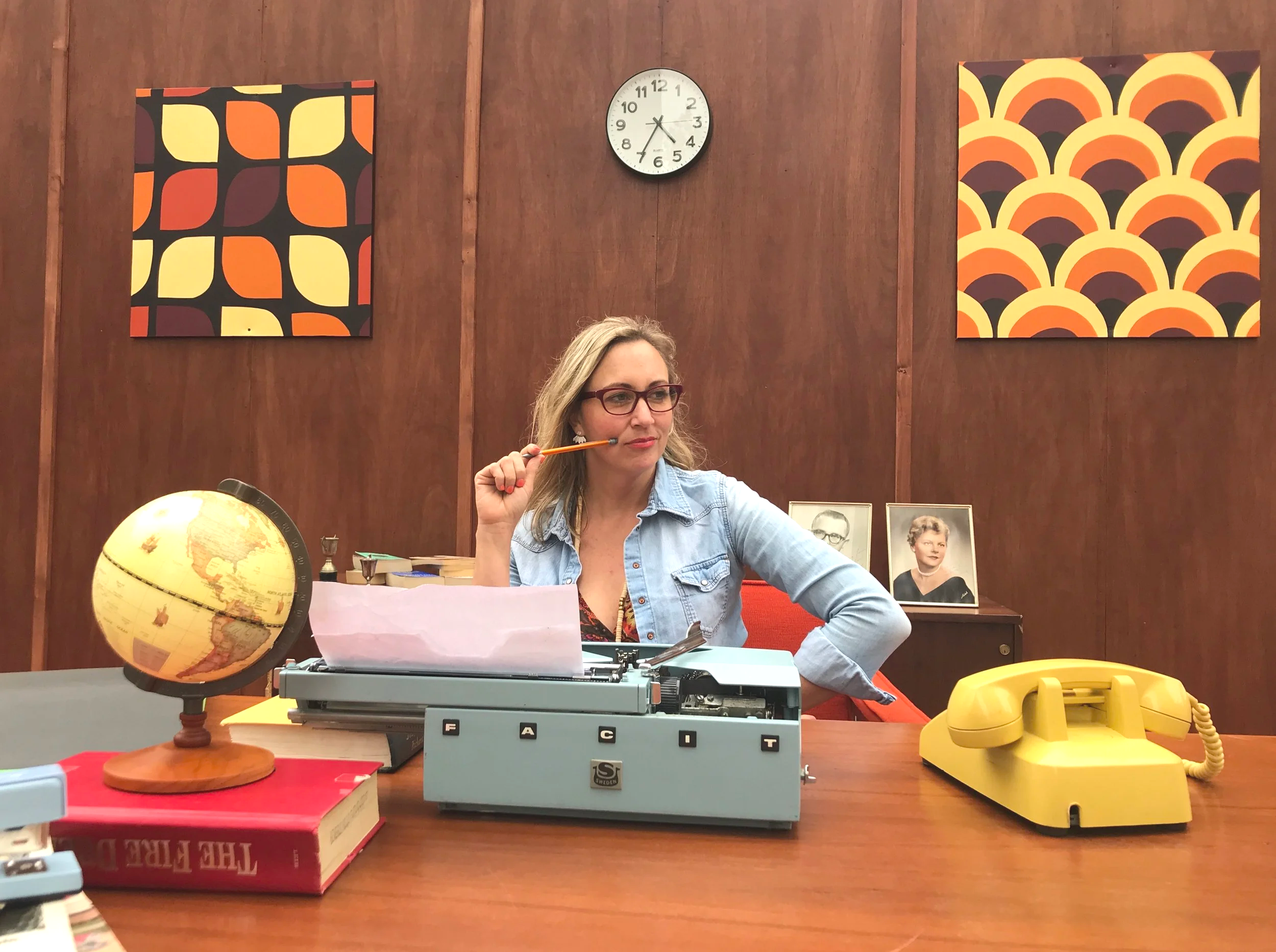 A woman sitting at a desk with vintage office items, a globe, framed photos, a typewriter, and a yellow rotary phone on a wooden table against a wooden wall with colorful abstract art and a clock in the background.