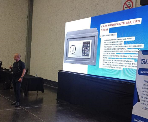 Pantalla grande con información sobre una caja fuerte de hotel en una presentación, acompañado de una persona con camiseta negra y gafas, en un ambiente interior con paredes grises.