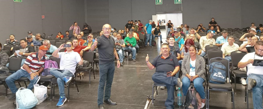 Grupo de personas en una sala, algunas usando dispositivos electrónicos, asistiendo a un evento o conferencia.
