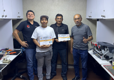 Cuatro personas en un taller, dos sosteniendo certificados y sonriendo, en un ambiente con herramientas y equipos tecnológicos.