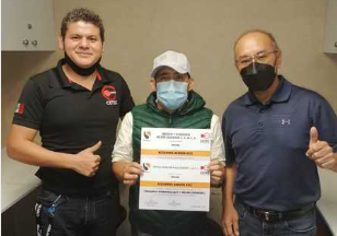 Tres hombres con mascarillas posando, uno sostiene dos certificados y todos muestran el pulgar hacia arriba.