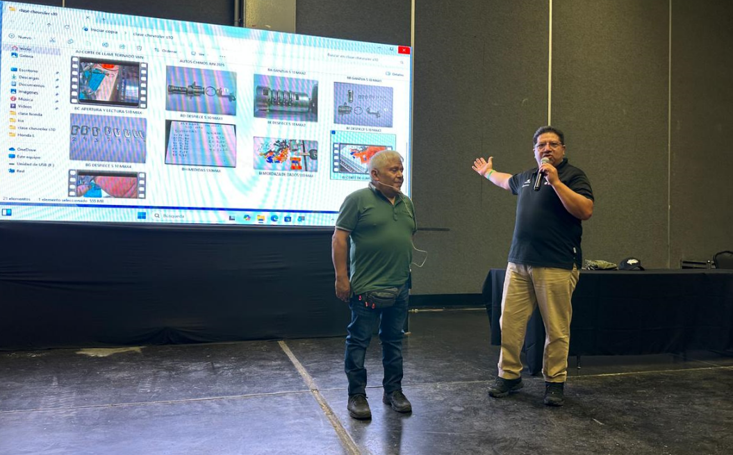 Dos hombres en una conferencia, uno con micrófono, frente a una pantalla grande con imágenes y documentos en una sala de conferencias.