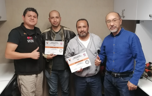 Cuatro hombres en un ambiente interior, dos de ellos sosteniendo certificados, uno con una mano en el pecho y otro con el pulgar hacia arriba.