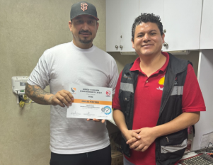 Dos hombres en una oficina, uno sosteniendo un cheque y el otro con mochila, ambos sonriendo.