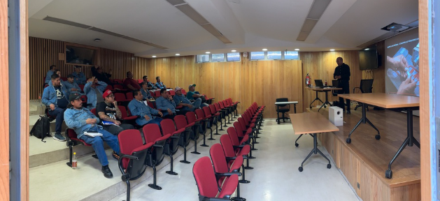 Salón de conferencias con varias personas sentadas en las butacas, asistiendo a una presentación en una pantalla grande, en un ambiente con paredes de madera y un escenario.