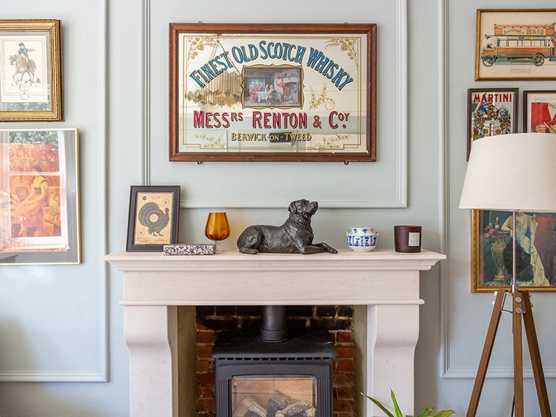 A cozy living room with a light gray fireplace decorated with eclectic items, including a vintage whisky ad, framed art, a lamp, a cast resin figurine urn of a Labrador, candles, and a potted plant.