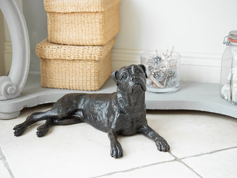 A realistic cast resin figurine Boxer urn lies on a tiled floor, with stacked woven baskets, a glass bowl of pine cones, and a jar on a curved gray surface.