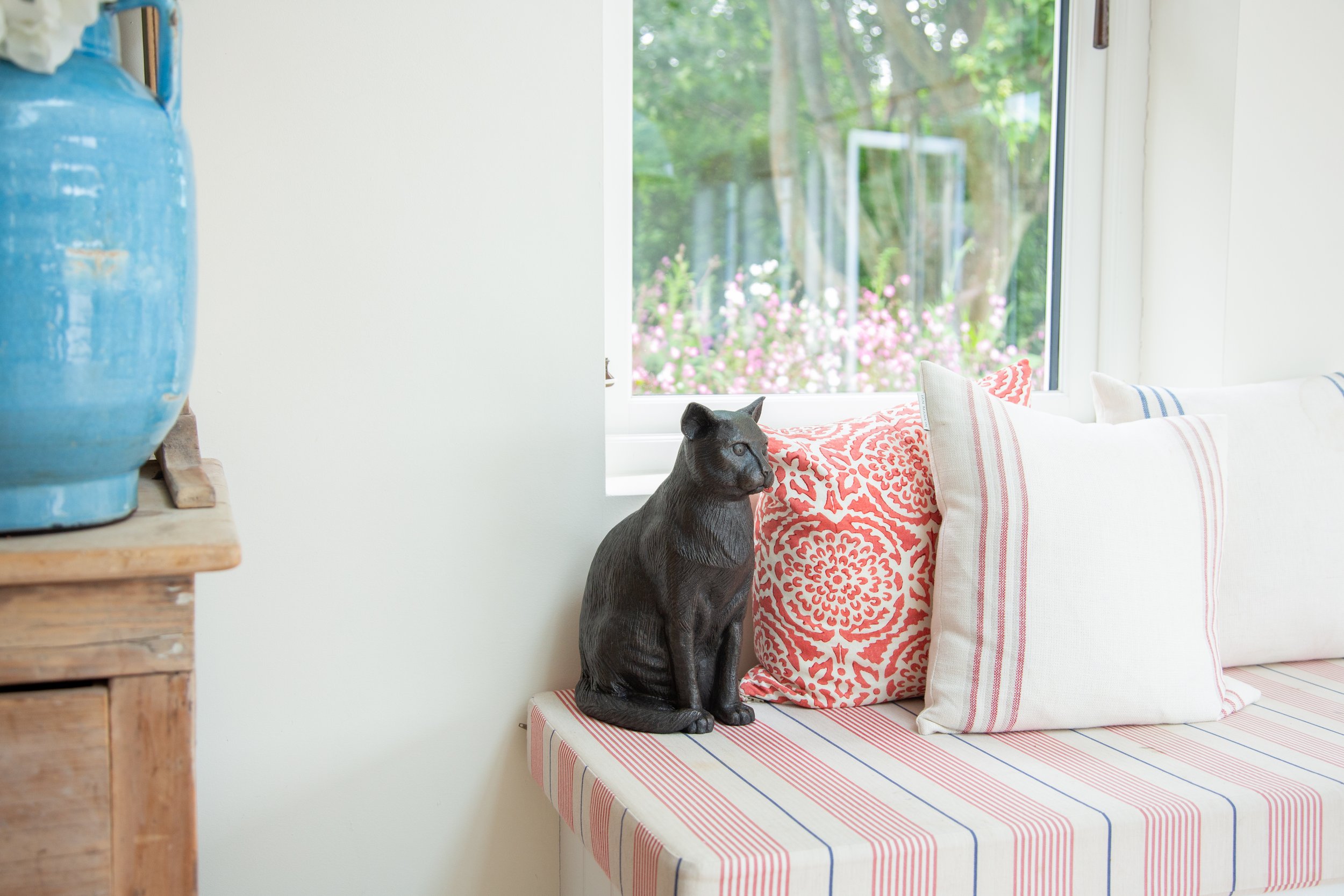 A figurine urn of a cast resin cat figurine urn with a bronze finish sits on a striped cushion bench beside decorative pillows and a blue vase, against a bright window view.