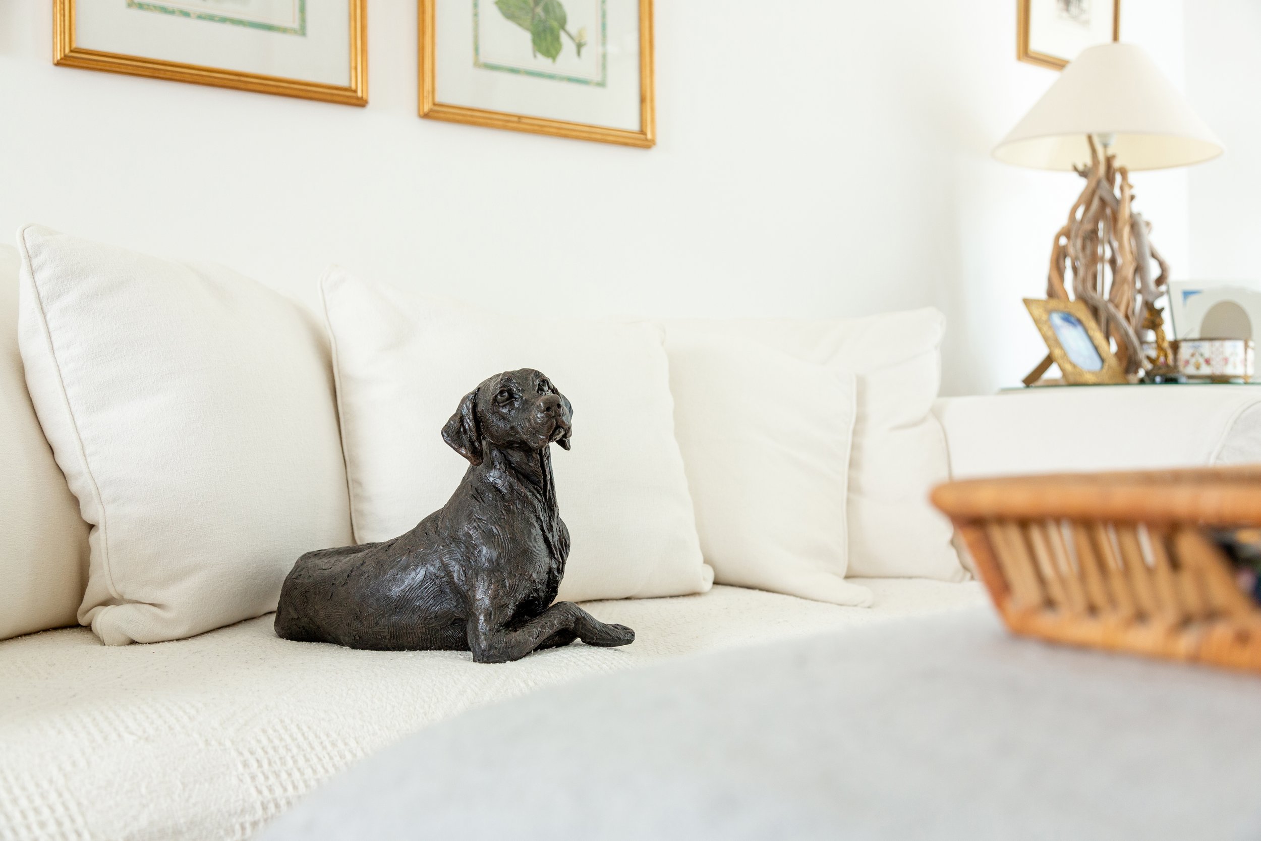 A cast resin figurine Pointer urn sits on a white couch with plush pillows, framed artwork on light walls, a lamp on a side table, and a basket in the foreground.