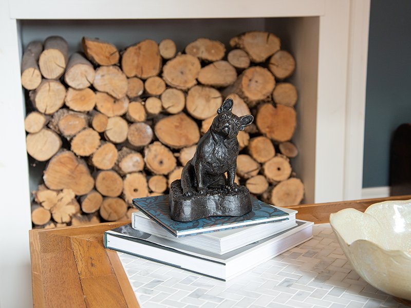 A cast resin figurine urn of a French Bulldog sits on stacked books on a wooden table, with a fireplace of cut logs behind it and a ceramic dish on a tiled surface to the right.