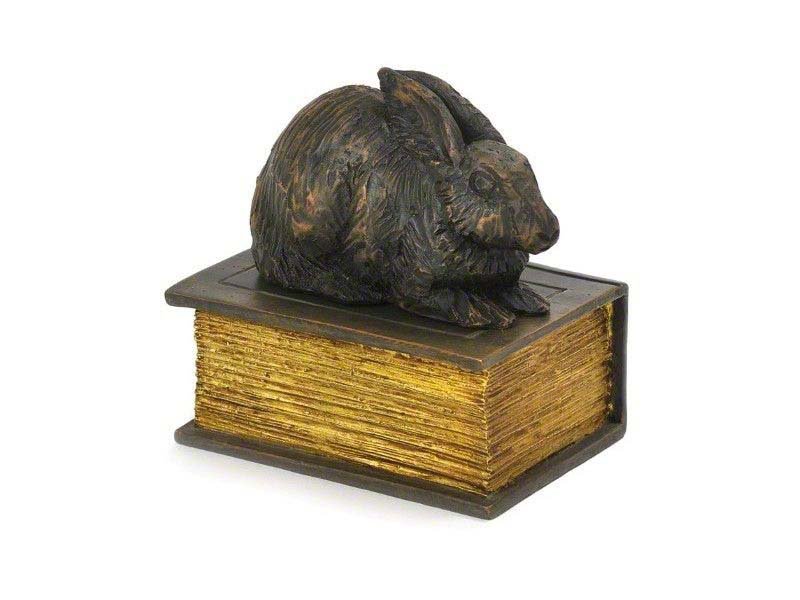 A cast resin rabbit figure urn on top of a closed book with gilded pages.