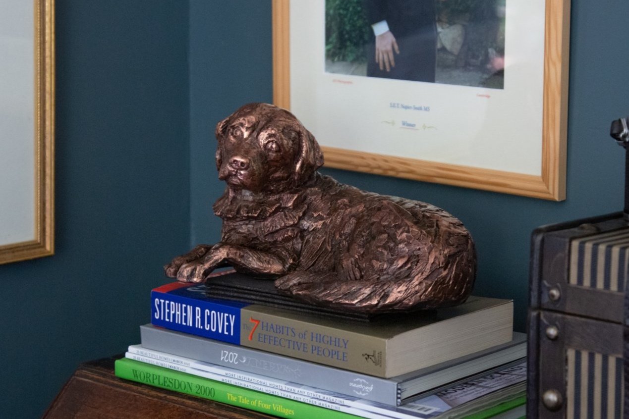 A copper plated Golden Retriever sculpture relaxes atop stacked books in a room with green walls and framed pictures in the background.