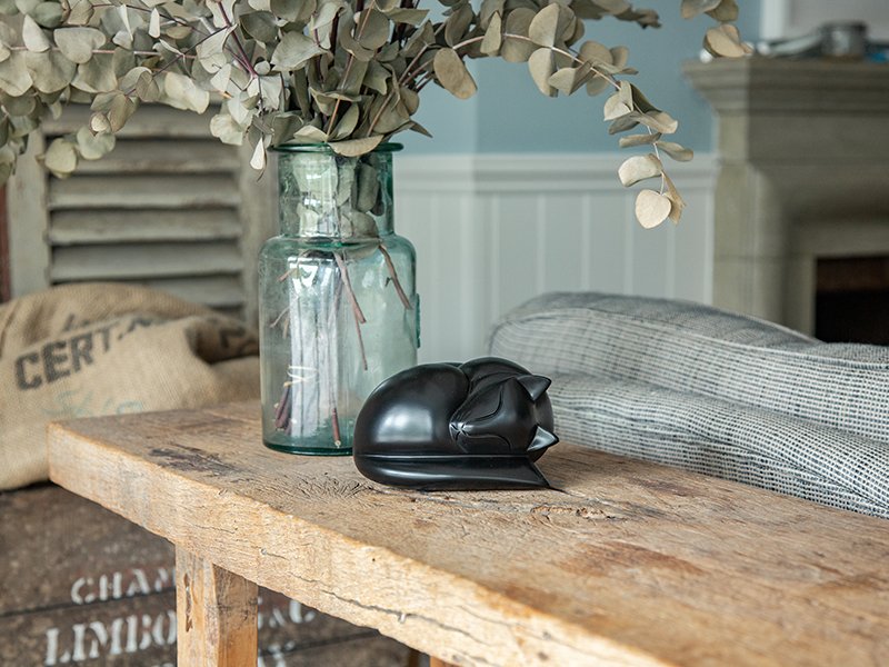 A cast resin black sleeping cat figurine urn on a wooden table beside a jar of eucalyptus, with rustic decor in the background.