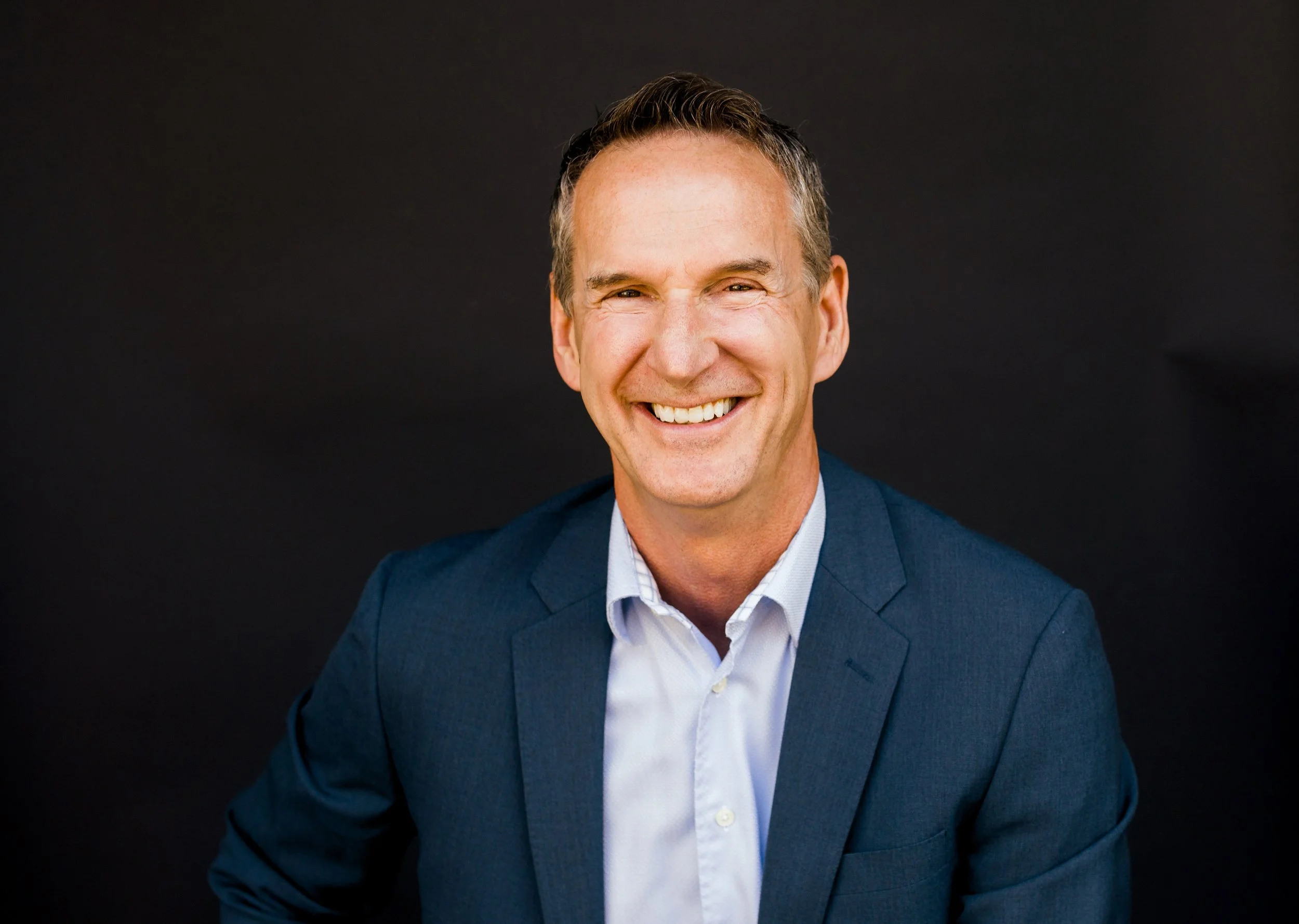 A smiling man in a blue suit and white dress shirt against a black background.